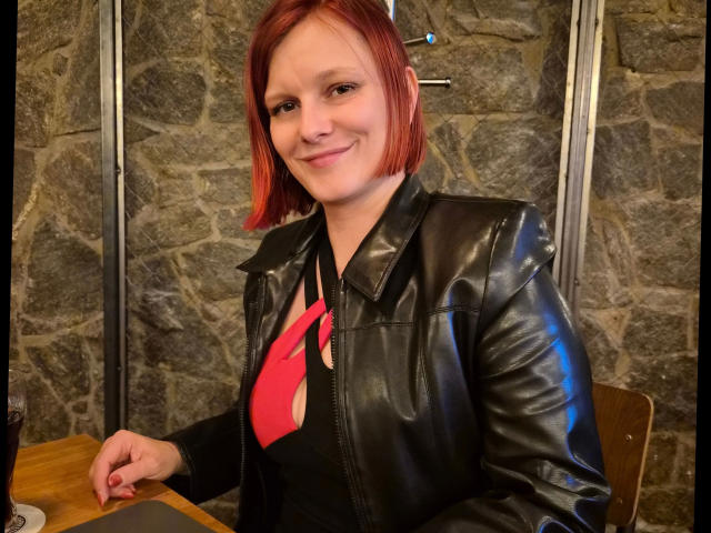 Ella-Bizarr, aantrekkelijk met kort rood haar, lachend, gekleed in een zwarte leren jas en een rode das, zittend aan een houten tafel. tegen een stenen muur, met een blik die seksuele spanning uitstraalt en die haar zelfvertrouwen weerspiegelt..