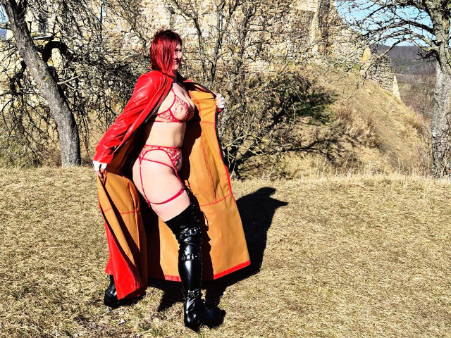 Ella-Bizarr-hot, une rousse séduisante, se tient dans un champ herbeux, vêtue de lingerie rouge, d'un long manteau rouge et de bottes noires. Son pénis est partiellement visible, ce qui crée une forte tension sexuelle. Couleurs vives et éclatantes.