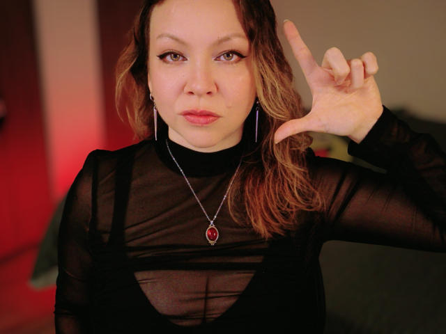 GoddessKrystalViolet, une femme à la peau pâle, aux cheveux bruns et aux yeux rouges, portant un haut noir transparent, un pendentif rouge et de l'argent. boucles d'oreilles, faisant un geste de la main comme pour dire "rock on", regard intense, fond rouge..