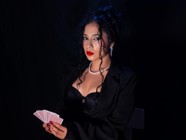 LilithStygian mit dunklen, lockigen Haaren, rotem Lippenstift, einem schwarzen Spitzen-BH und einem schwarzen Blazer, hält Pokerkarten und trägt eine Perlenkette. Kette. Schwarzer Hintergrund. Verführerischer, intensiver Blick, sexy Ausstrahlung..