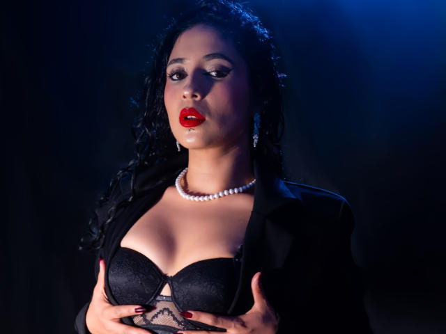 LilithStygian, a dark-haired woman with wet hair, wears red lipstick, a pearl necklace, and a black lace bra with matching black blazer, gripping her breasts. Blue lighting highlights her face.