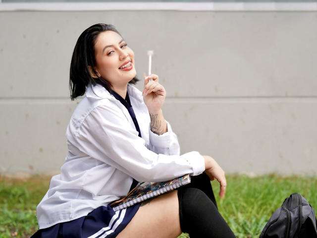 AmyRoss sits on grass, smiling, holding a lollipop near her lips. She wears a white shirt, black skirt, and thigh-high socks. Her black hair contrasts with the bright background.