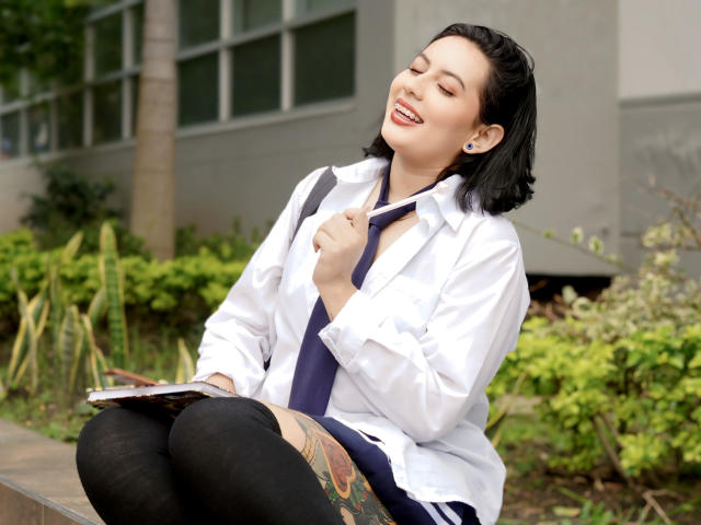 AmyRoss with short black hair, wearing a white shirt and black tie, sitting with legs crossed, black leggings, colorful thigh tattoo, smiling, eyes closed, sunlight, green plants in background.