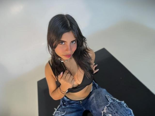 Photographie de Celeste Morel, aux cheveux foncés, regardant vers le haut avec un regard séduisant, portant un crop top noir et un jean déchiré. debout devant une table noire, regardant directement la caméra.