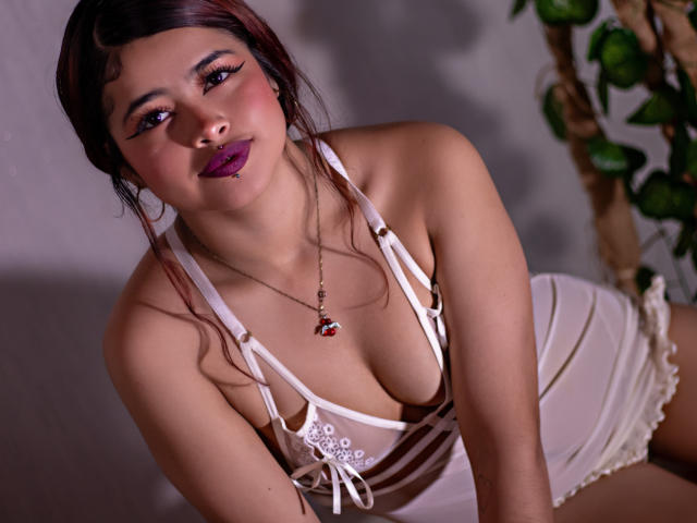 JennieRider, kurvig, oben ohne, weiße Dessous, tiefer Ausschnitt, braunes Haar, lila Lippenstift, verführerischer Blick, angelehnt vorwärts gerichtet, Umriss der Brustwarze, Halskette mit rotem Stein, Topfpflanze im Hintergrund, weiches Licht, sexuelle Spannung.