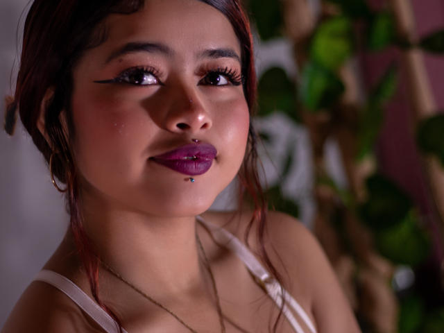 Fotografia ravvicinata di JennieRider, una donna dai capelli scuri con rossetto viola lucido, labbro inferiore con piercing e eyeliner nero. Sta guardando di lato con un'espressione leggermente seducente. Pianta verde sfocata sullo sfondo.
