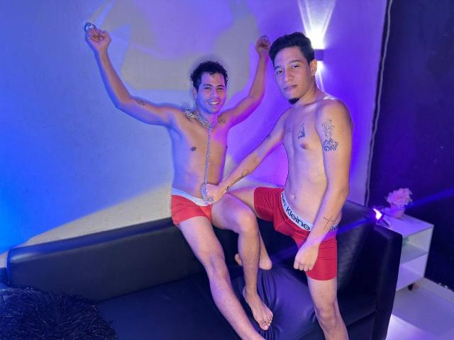 Two shirtless MaikolAndAndry guys with short black hair, wearing red shorts, sitting on a black couch. Blue and purple lighting, one raising arm, other holding hands. Visible tattoos, smiling.