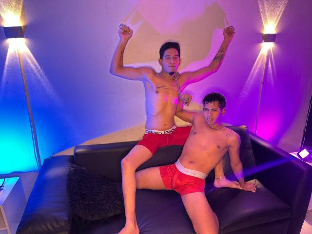 Twee gespierde mannen in rode boxershorts, MaikolAndAndry, staan met armen omhoog, terwijl de andere MaikolAndAndry op een zwarte ondergrond zit. bank, neonverlichting, seksuele spanning, tatoeages, donker haar, slanke lichamen.