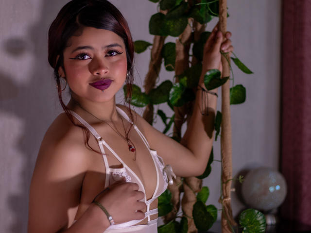 JennieRider, Asian woman, dark hair, purple lipstick, white lace bra, holding plant, shadow on her face, confident gaze, seductive pose, necklace with red pendant, dimly lit, sexy vibe, soft shadows.