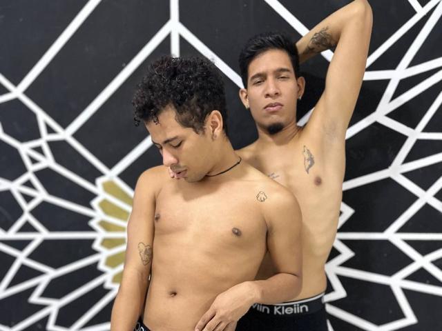 Two shirtless, tan-skinned men with tattoos, MaikolAndAndry in front, arm raised, cock visible, black hair, MaikolAndAndry in back, arm around, relaxed, confident, black and white geometric background.
