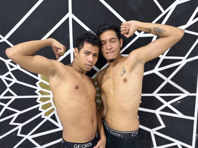 Dos hombres musculosos, MaikolAndAndry, sin camisa, mostrando sus bíceps, de pie muy cerca el uno del otro. Ambos tienen el cabello oscuro, tatuajes visibles y llevan ropa interior negra con las etiquetas blancas "GECKO". Fondo geométrico en blanco y negro. Tensión sexual.
