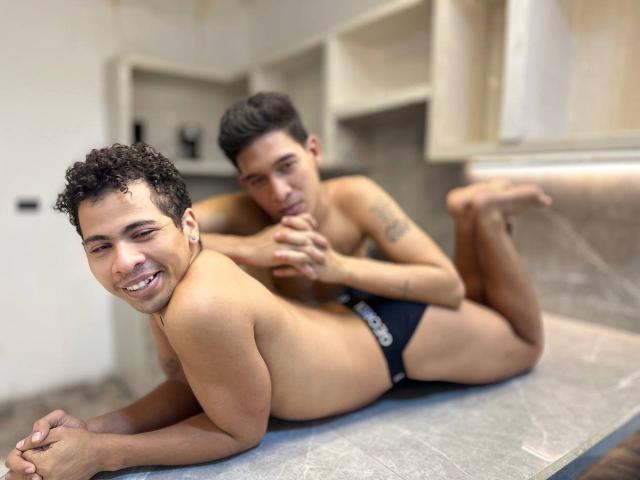 Dois jovens sem camisa, com cabelos pretos encaracolados e pele clara, deitados em uma bancada de cozinha. MaikolAndAndry na frente, sorrindo, usando cuecas pretas. Segundo MaikolAndAndry atrás, chupando os dedos. Tensão sexual.