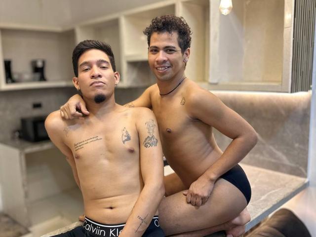 Dos hombres sin camisa, tatuados y con cabello corto y rizado, MaikolAndAndry a la izquierda y MaikolAndAndry a la derecha, ambos vestidos de negro. ropa interior, arrodillados juntos, MaikolAndAndry a la derecha tiene la mano en el hombro de MaikolAndAndry.