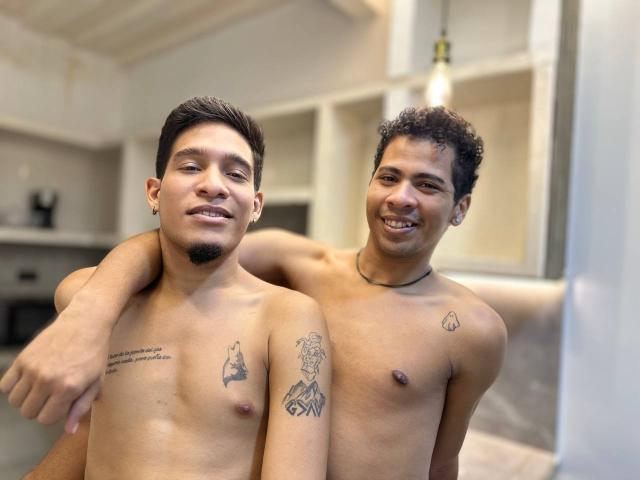 Two shirtless young men with medium brown skin and short curly hair, standing close, one arm around the other. Tattoos on chests. Bright room, hanging light bulb. Smiling, relaxed.