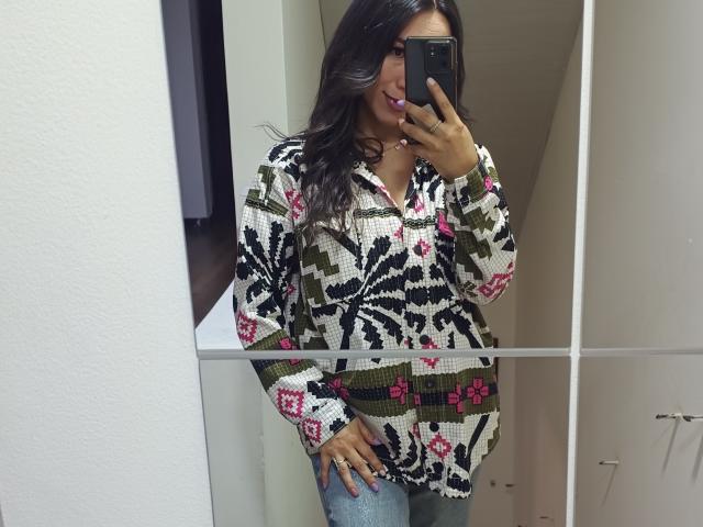 MiaReed taking a mirror selfie, holding a black phone in her right hand. She's wearing a colorful, patterned sweater with pink and green flowers, and tight blue jeans. Her long black hair is slightly wavy.