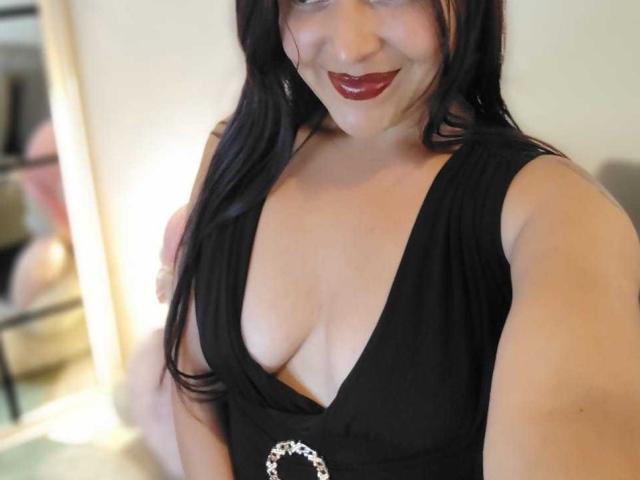 Close-up de AfroditaLaDiosa, uma mulher curvilínea com cabelos longos e pretos, vestindo um vestido preto com decote baixo que revela o colo. batom vermelho, sorriso sedutor, luz suave, fundo ligeiramente desfocado..