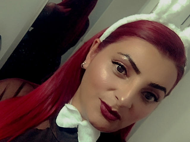 Close-up foto van SharaLovee met felrood haar, die een witte hoofdband met konijnenoren en een strik draagt. Ze heeft donkere lippenstift, dikke wenkbrauwen en kijkt recht in de camera met een enigszins verleidelijke uitdrukking..