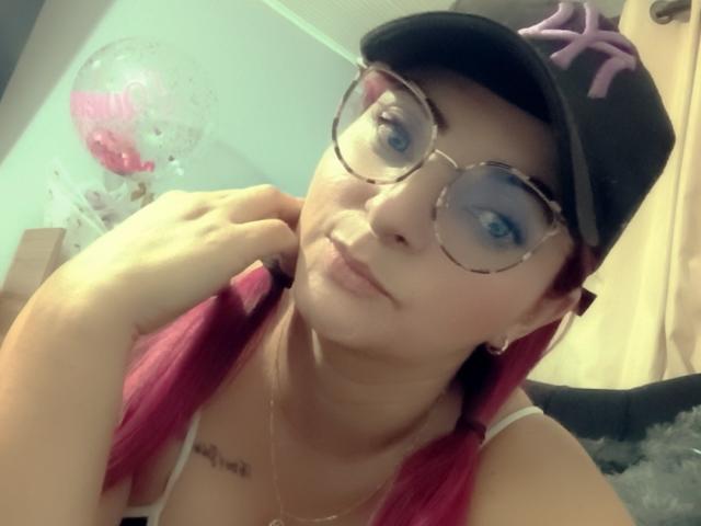 Foto ravvicinata di SharaLovee, con la pelle chiara e i capelli rossi, che indossa un berretto nero con una stella viola, occhiali rotondi, bianchi. un canottino e una collana. Lei sta guardando la fotocamera, con la mano sul mento..