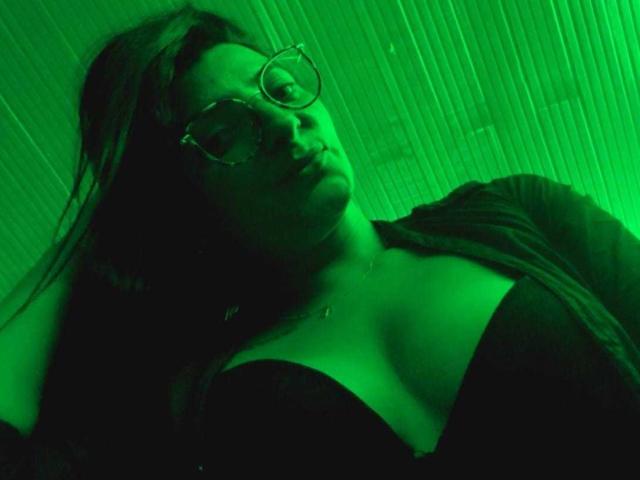 Photographie prise en contre-plongée de SharaLovee, portant des lunettes et un haut noir à décolleté plongeant, éclairée par une lumière néon verte. Longs cheveux noirs, expression sérieuse, un éclat vert illumine son visage et sa poitrine..