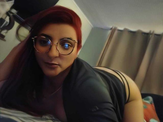 SharaLovee, met rood haar en een bril, leunt voorover op een bed, gekleed in een zwarte top en een zwarte string, waarbij haar achterwerk zichtbaar is. Ze kijkt met een lichte glimlach naar de camera. Slaapkamer setting..