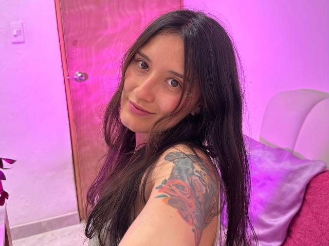 KimyRosse, con cabello negro largo, ojos marrones y un tatuaje en el hombro derecho, se toma un selfie. Ella está mirando a la cámara con una sonrisa sutil, en una habitación iluminada con luz rosa. Increíblemente atractivo/a..