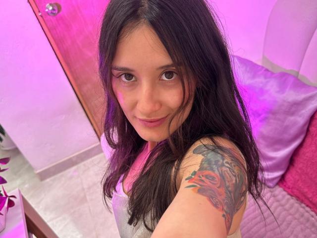 Selfie de primer plano de KimyRosse, una joven con cabello largo y negro, ojos marrones y un tatuaje colorido en su lado derecho. hombro.

ella lleva una blusa blanca y mira directamente a la cámara con una sonrisa sutil. Iluminación de color rosa brillante y púrpura en el fondo.