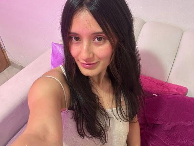 KimyRosse mit langen, schwarzen Haaren, die leicht lächelt, trägt ein weißes Top mit Spaghetti-Trägern und streckt den Arm für ein Selfie aus, rosa. und violette Kissen auf einer weißen Couch, sanfte rosa Beleuchtung. Total süß..