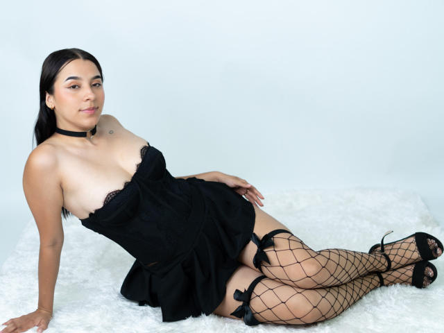 Sammy Rodriguez, avec ses cheveux foncés, allongée sur une fourrure blanche, portant une robe noire en dentelle sans bretelles, des bas résille et du noir talons hauts, qui dégagent une tension sexuelle. Collier noir autour du cou. Pose séduisante et confiante..