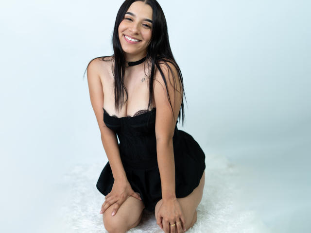 Sammy Rodriguez est agenouillée sur une surface blanche, vêtue d'une robe noire sans bretelles, souriante, avec de longs cheveux noirs, les mains posées sur ses genoux. corps mince, peau claire, petites seins, collier noir, fond blanc.