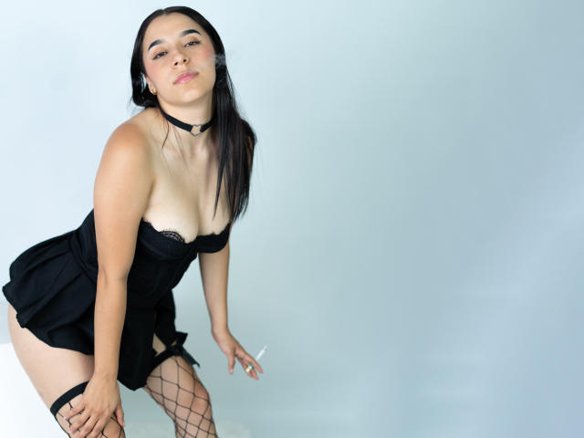 Sammy Rodriguez, penchée en avant avec un air boudeur, portant un haut en dentelle noire, une jupe noire courte, des bas résille et du noir collier ras du cou. Peau pâle, longs cheveux noirs, attitude provocante, pose séduisante..