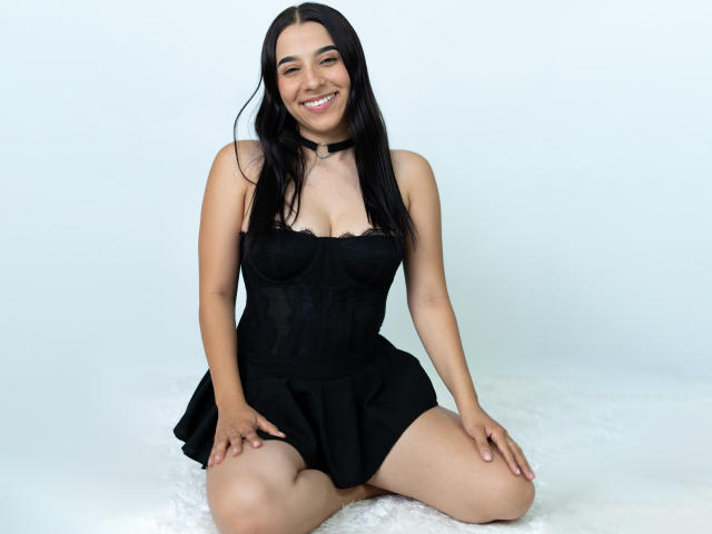 SammyRodriguez, arrodillada con las piernas separadas, vestida con un vestido negro sin tirantes, sonriendo, con cabello negro largo, piel pálida, de raza blanca. fondo, manos apoyadas en los muslos, iluminación suave, tensión sexual, ligera sombra debajo del vestido..