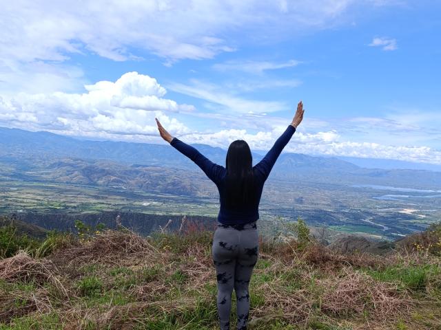 PauletteLoveX está de braços levantados, olhando para uma vasta paisagem. Ela veste uma jaqueta escura e calças claras, com um céu azul brilhante ao fundo, com algumas nuvens espalhadas. Grama marrom e levemente desfocada em primeiro plano..
