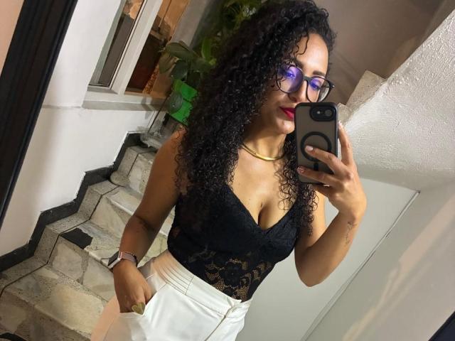 MoonBlackSs, uma mulher curvilínea com longos cabelos cacheados pretos, usando óculos, uma blusa de renda preta e calças brancas, tirando uma foto no espelho. selfie. Ela tem uma expressão sensual e confiante, com um toque de sedução..