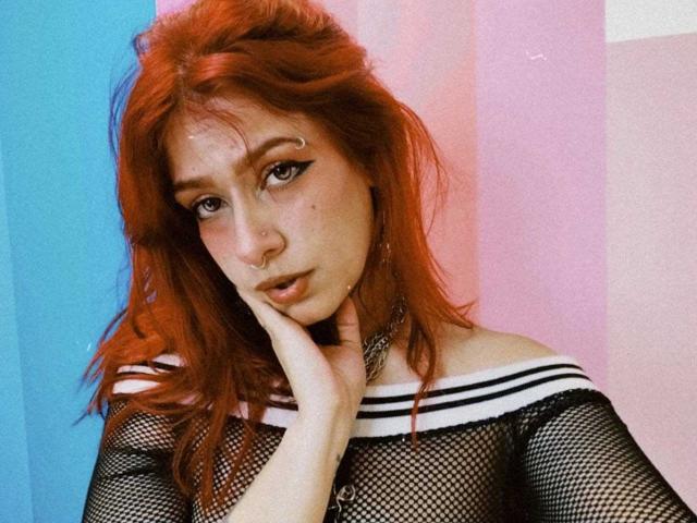 Fotografia di LolyYunno con i capelli rossi, che indossa una maglia nera a rete con strisce bianche, con una mano che tocca il viso, sfondo blu e sfondo rosa, illuminazione soffusa, espressione seducente, guance leggermente arrossate.