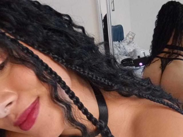 Close-up photo of EricaRousex with curly black braids covering her face, red lipstick, black strap choker, partial reflection of her back in the mirror, tense sexual vibe, grey background.