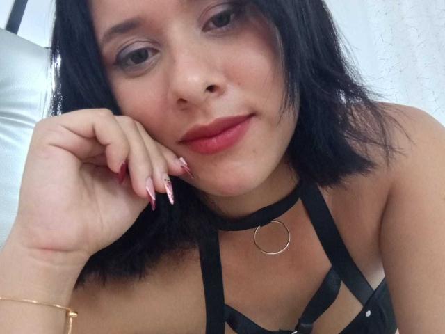 Primer plano de EricaRousex, una mujer de piel clara con cabello negro, lápiz labial rojo y un top negro con tirantes. Ella apoya su rostro en su mano, mostrando uñas largas y rosadas, un anillo de oro y un collar negro. Increíblemente atractivo/a..