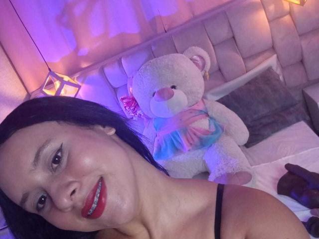 Photograph of EricaRousex, close-up, smiling with red lipstick, black hair, holding a white teddy bear with blue ribbon, soft purple and pink lighting, bed with tufted headboard in background.