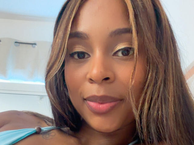 Close-up photo of EmmaLatin with brown and blonde streaked hair, brown eyes, and pink lips. She's wearing a blue strap, looking directly at the camera with a subtle smile. Bright natural light.