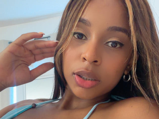 Close-up photo of EmmaLatin, a young Black woman with light brown skin and straight brown hair, looking directly at the camera with slightly parted pink lips. She wears a blue strap top. Her hand touches her cheek.