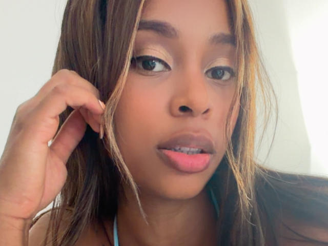 Close-up foto van EmmaLatin met bruin haar, lichtbruine huid en roze lippenstift, die recht in de camera kijkt. Lichte glimlach, hand die haar oor aanraakt, heldere achtergrond. Heel aantrekkelijk..