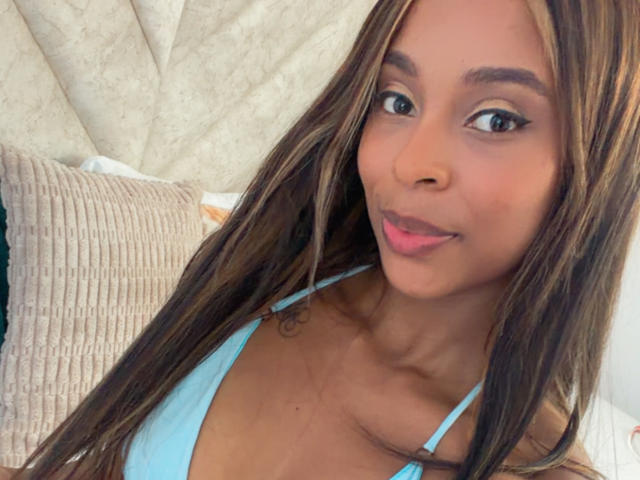 Close-up of EmmaLatin, a sexy woman with long brown hair, wearing a light blue tank top, smiling with pink lips. Her eyes are large and dark, and she's leaning slightly forward.