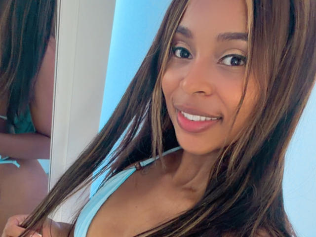 Selfie de perto de EmmaLatin, com cabelos castanhos longos, sorrindo, vestindo uma regata azul clara, mostrando um pouco de sua grande seios e braços tonificados, fundo azul claro, reflexo no espelho..