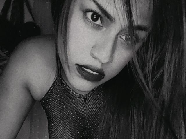 Photographie en gros plan d'IvannaDuran, portant un haut en maille, avec du rouge à lèvres foncé, un regard intense et un visage partiellement dissimulé. Noir et blanc, contraste élevé, évoquant une tension sexuelle. Cheveux foncés, arrière-plan sombre..