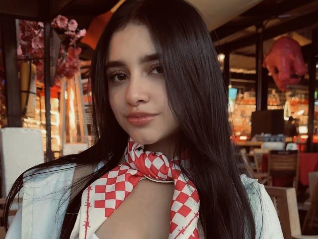MeganPaige, a hot woman with long black hair, wearing a red and white checkered scarf, looking directly at the camera, slightly open white shirt showing cleavage, sexy vibe, dimly lit cafe background.