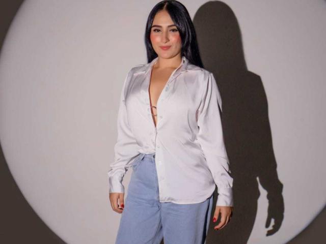 HiilaryDolce stands against a white wall, shadowed to the right. She wears an unbuttoned white shirt revealing cleavage and light blue high-waisted pants. Shadowed cock visible through shirt.