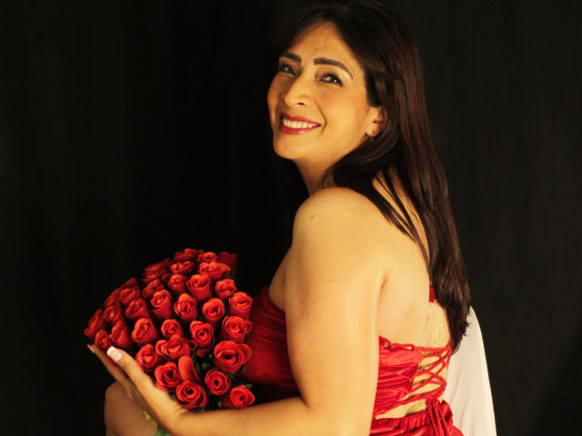 DolceMartinni with long dark hair, smiling at the camera, holding a bouquet of red roses. She's wearing a strapless red dress, black background, bright red roses contrasting with her tan skin.