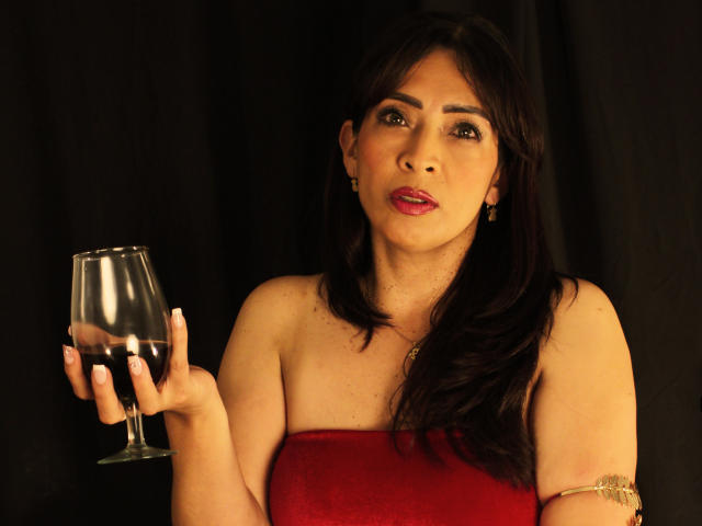 DolceMartinni, con i capelli scuri e un abito rosso senza spalline, tiene un bicchiere di vino rosso nella mano destra. Ha il rossetto rosso, una fascia al braccio e uno sfondo nero. Incredibilmente sexy..