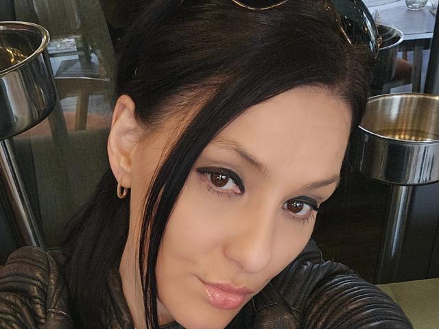 Close-up selfie of JoliDanielle, an Asian woman with fair skin, black hair, and brown eyes. She has a slight smile, glossy pink lips, wearing a black top, and small earrings. Shiny lamps in background. Sexual tension from direct gaze and close-up framing.