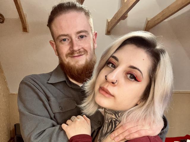 Fotografie von AllisonXAlfred, einem Mann mit Bart und einem grauen Hemd, und einer Frau mit platinblondem Haar, Nasenring und roter Kleidung. Lidschatten. Er lächelt, sie schaut in die Kamera, ihre Hand liegt auf seiner Schulter..