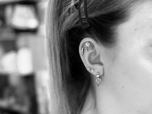 Fotografía en blanco y negro de la oreja de SugarrX, con tres pendientes de diamantes, y el cabello recogido con una pinza. Fondo con luz difuminada. Enfoque en la oreja y los pendientes, con una insinuación de tensión sexual..